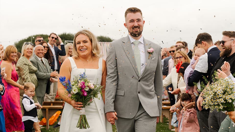 A close up of Sian in her white wedding dress and Danny in his grey suit walking down the aisle in between friends and family who are all smiling.